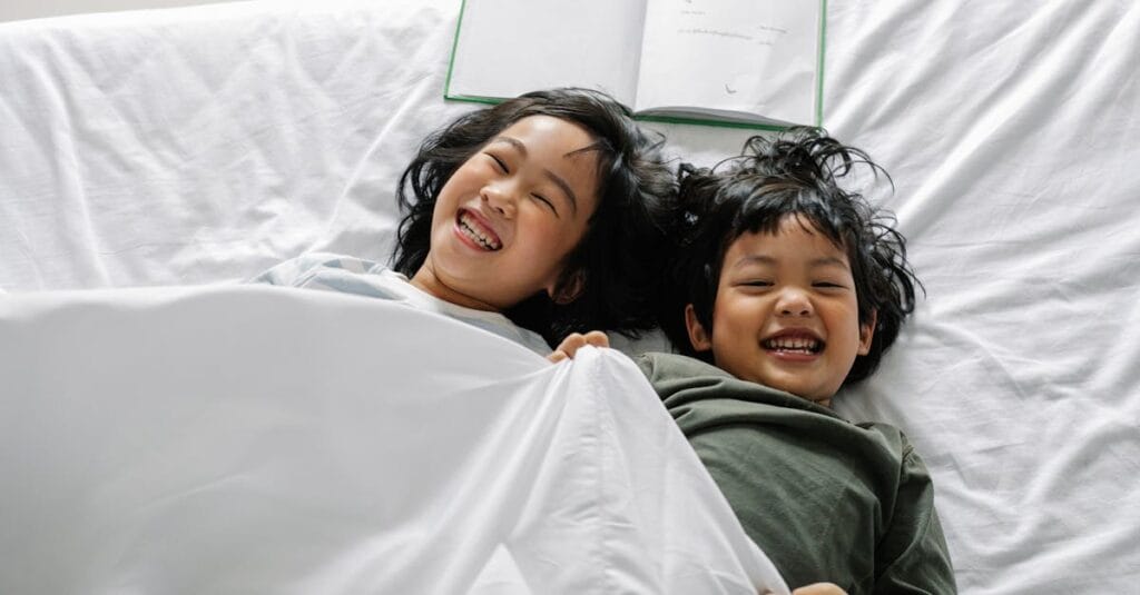 From above of Asian siblings laughing happily while lying in bed under soft blanket